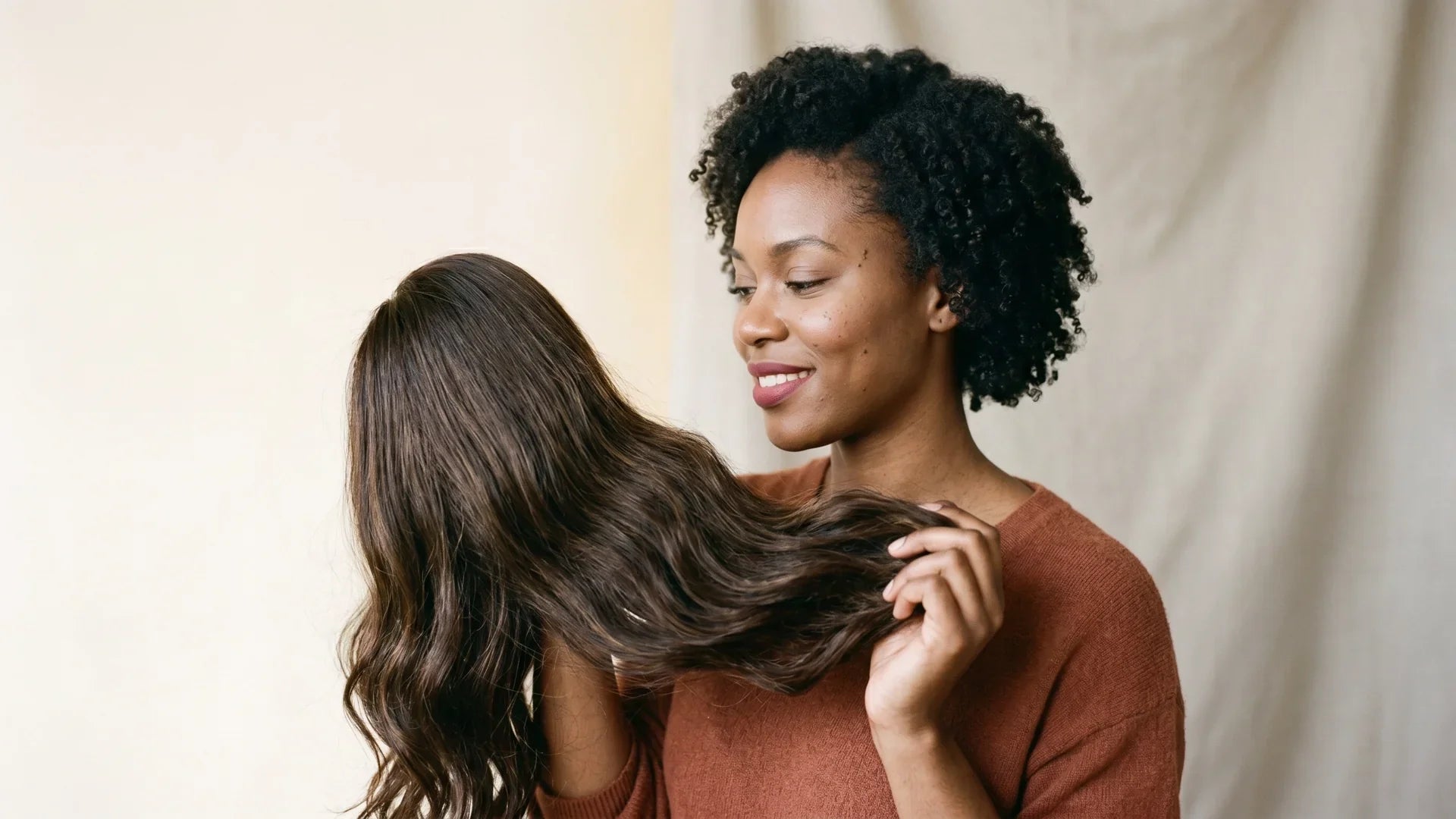 A smiling woman with short natural curls admiring a long wavy Mimicing glueless wig before installation.