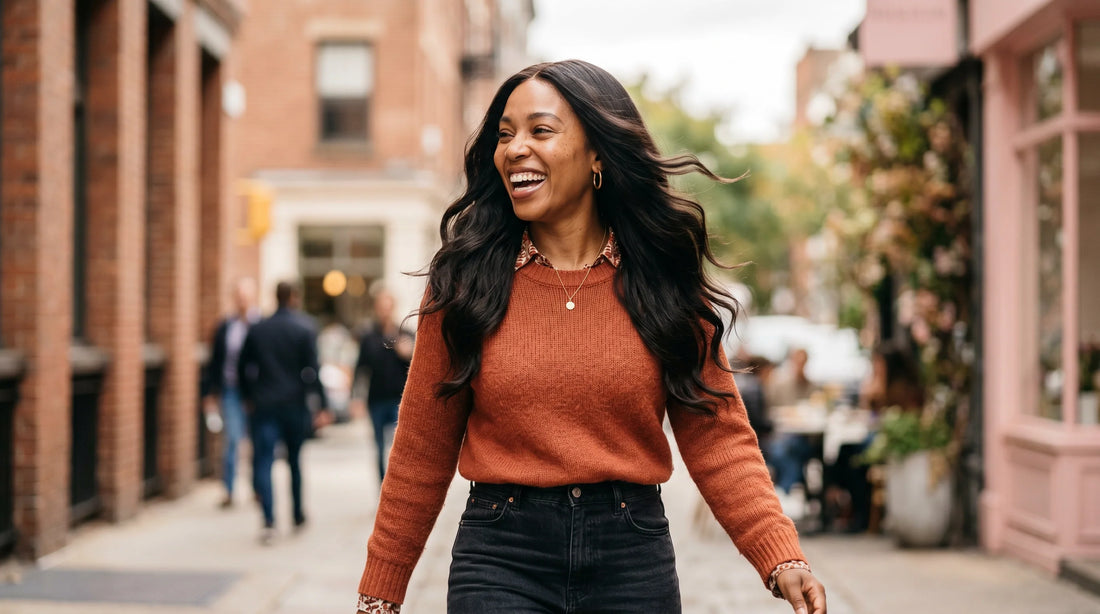 A confident, smiling woman walking down a city street with her long wavy wear-and-go skin lace wig flowing naturally in the wind, showcasing a secure glueless fit.