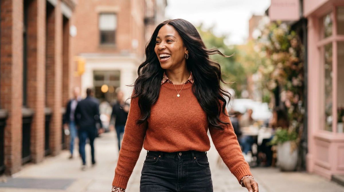A confident, smiling woman walking down a city street with her long wavy wear-and-go skin lace wig flowing naturally in the wind, showcasing a secure glueless fit.